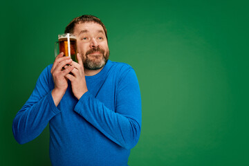 Portrait of emotive man in blue sweater posing with beer glass isolated on green background. Delightful, relaxed look