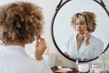 Young woman doing massage of face and spreading applied serum on skin with quartz roller while...
