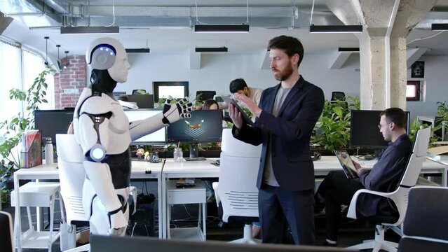 Side-view of young professional IT worker programming white futuristic robot with artificial intelligence using tablet. Scientific cyborg moving arms and hands standing in office. AI concept.