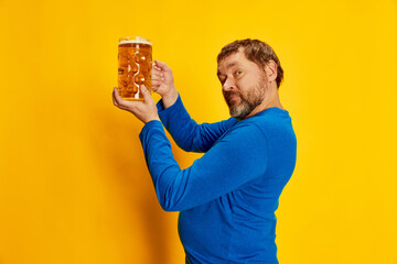 Portrait of emotive man in blue sweater posing with foamy beer mug isolated over yellow background....