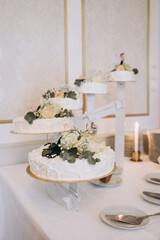 Wedding. Banquet. A cake for the bride and groom stands on a table with a white tablecloth, candles stand nearby and is decorated with flowers and greenery. Nearby are candles, plates and cutlery