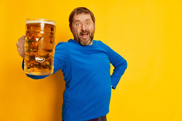 Portrait of emotive man in blue sweater cheerfully posing with foamy beer mug isolated over yellow...