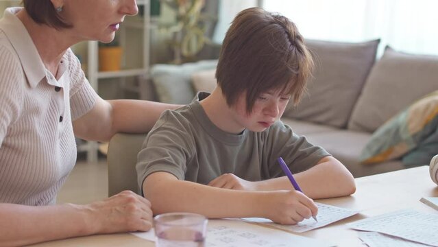 Caring mother helping her 14 year old daughter with down syndrome with school homework, sitting together at desk in living room