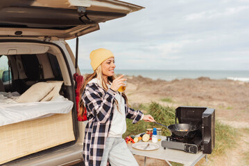 happy nomad woman having glass of beer while cooking outside camper van next to the beach. Concept...