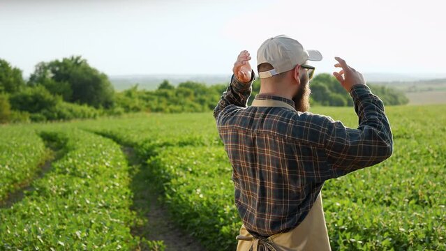 Back view of young farmer standing on field, putting on cap. Male with beard wearing plaid shirt and apron, looking on plants, examining. Concept of working in countryside.
