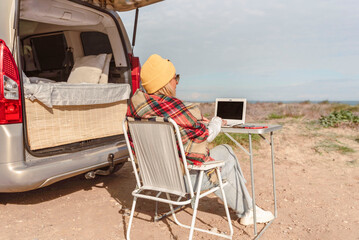Adult woman works on laptop while traveling with camper van. Concept of modern people lifestyle in smart working or digital nomad freedom
