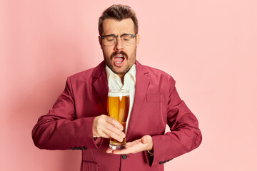 Portrait of stylish emotive man in a suit posing with glass of lager beer isolated on pink background. Traditional hop taste