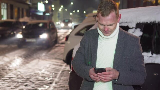 Handsome young man using smartphone on crowded street outdoors under snowfall near passing by car at night. Checks mail, chats or news on Internet. Posting social media, online shopping, texting, e