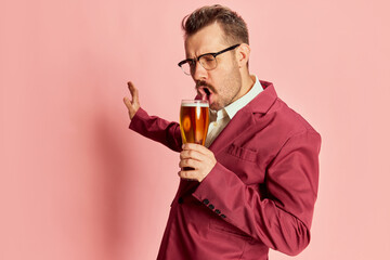 Portrait of stylish emotive man in a suit posing with glass of lager beer isolated on pink...