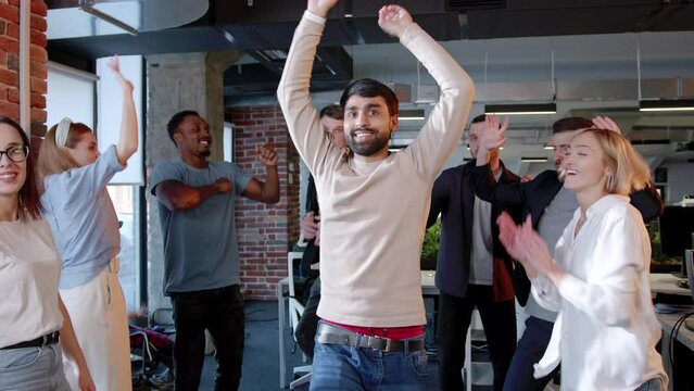 Happy young successful workers of company dancing having fun on corporate party in office. Multiethnic group of people employees with Indian man in center smiling moving celebrating success.