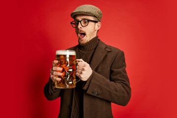 Portrait of stylish man in classical clothes posing with glass of foamy lager beer isolated on red background. Drinking with excitement