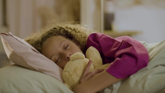 Cute young girl sleeping in bed holding soft toy. Pretty blonde child in pink pyjamas hugging teddy bear, enjoying deep sleep. Slider closeup shot. Childhood, sleep concept.