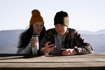 Couple with thermos is sitting in front of mountains view and looking in a phone