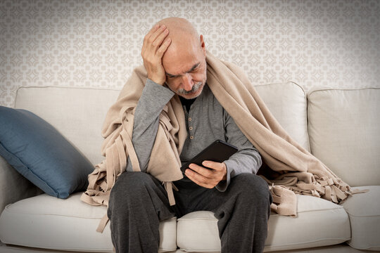 Serious Sad Old Man Sitting with Blanket on Shoulders with Mobile Phone in Hand