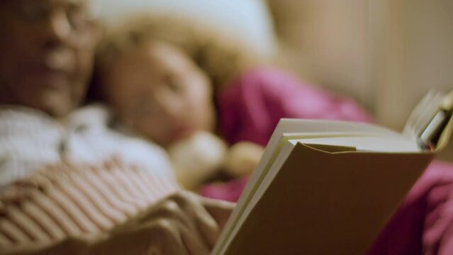Ederly man reading goodnight fairy tale for his granddaughter. Closeup of male hand turning page. Camera focus on book in foreground. Blurred silhouettes in background. Parenthood, leisure concept.