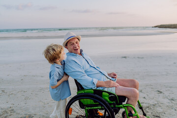 Little boy pushing his granfather on wheelchair, enjoying sea together.