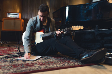 full shot of a young guitarist man sitting on the floor with a bass guitar and writing notes in the...