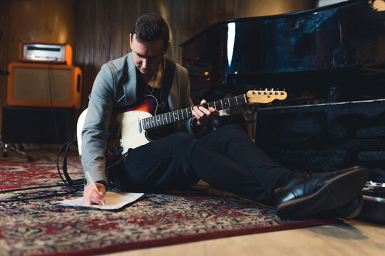 full shot of a young guitarist man sitting on the floor with a bass guitar and writing notes in the studio. High quality photo