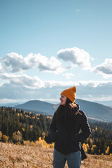 Young happy woman in orange beanie is standing in front of mountains landscape