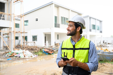 portrait of young engineer in vest with white helmet standing on construction site, smiling and...