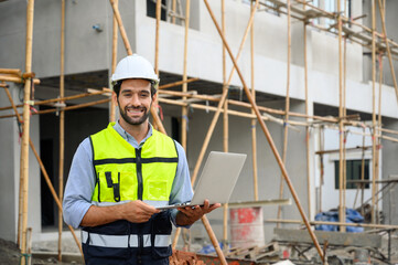 Young attractive construction smiling bearded in vest with white helmet working with laptop, standing on building construction site. Home building project.