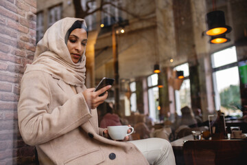 Young muslim woman with smartphone enjoying cup of coffe in cafe.