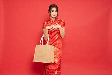 Fototapeta na wymiar Happy Asian woman wearing red traditional cheongsam qipao dress holding textile eco bag isolated on red background, Sustainable and environment protection concept