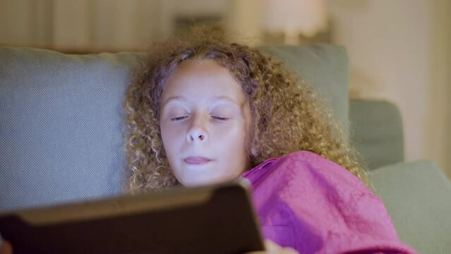 Pretty teenaged girl reading ebook before falling asleep. Young blonde lady lying in bed, holding digital tablet. Screen light reflecting on her face. Closeup shot. Children, modern technology concept