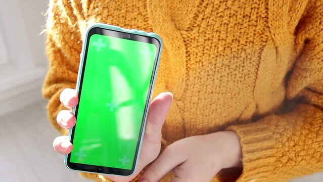 A girl in a yellow sweater uses a smartphone in a blue case and flips to the camera with a green screen. The concept of the energy and economic crisis.