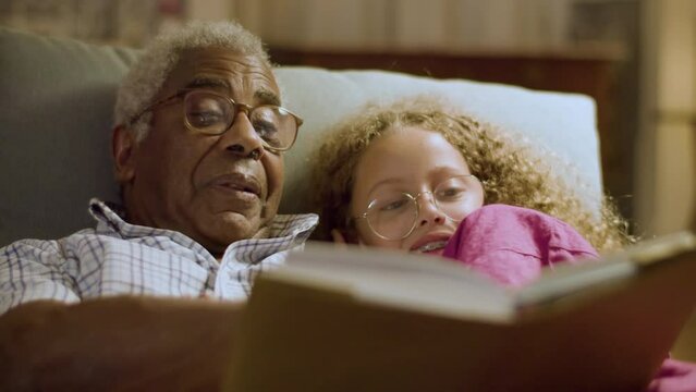 Cute girl and her grandpa lying on couch reading book together before going to bed. Elderly grey-haired man turning page while granddaughter listening and smiling. Family bonding, parenthood concept.
