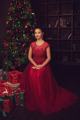 Beautiful portrait of a young girl in a red ball gown with a festive make-up on the background of a Christmas tree with a garland and boxes with gifts, New Year