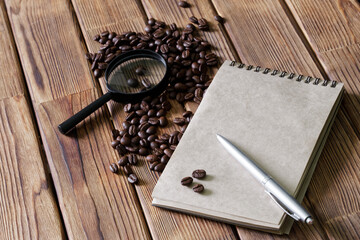 Obraz na płótnie Canvas Roasted coffee beans, notepad, magnifier and pen on a pine plank table. Concept of research and examination of coffee quality. Content of useful substances. Pre-sale preparation.
