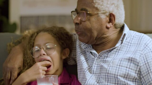 Closeup of young girl and granddad having late snack on couch. Cute blonde mixed race girl dipping biscuit in milk and biting it. Grandpa kissing granddaughters forehead gently. Happy family concept. 