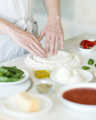 Obraz na płótnie Canvas Process of making pizza. Cook kneads dough with his hands on table. Ingredients for dough on table. Light background. View from above. Soft focus.