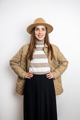 Smiling young woman in a jacket in a vu hat midi skirt against a white wall.