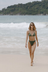 Woman in green bikini walking on beach. Against the background of waves. In sunglasses High quality photo