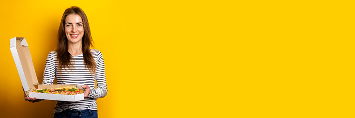 smiling young woman holding package with hot fresh pizza on yellow background. Banner