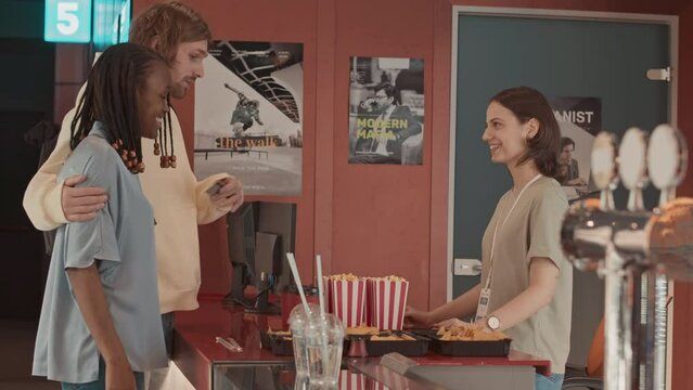 Young interracial couple talking to cinema girl while buying nachos at counter on date night