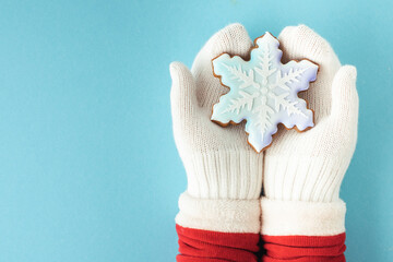 Woman hands wearing white mittens and holding snowflake shaped gingerbread cookie with festive icing on the blue pastel background. Christmas background