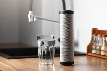 A glass of clean fresh water and carbon filter cartridge on wooden table in a kitchen interior....
