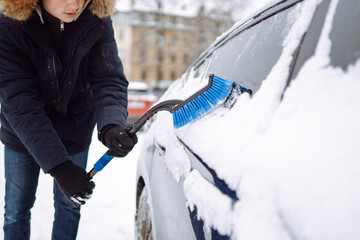 Portrait of young man cleaning snow off  car during winter snowfall. Transport, winter, weather, people and vehicle concept.