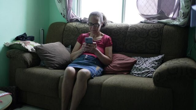 One candid hispanic senior woman using phone sitting at home couch. A casual older person looking at smartphone screen