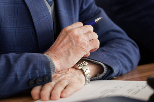 An adult man in a warm jacket holds a ballpoint pen in his hands, studying a paper document. Selective focus.