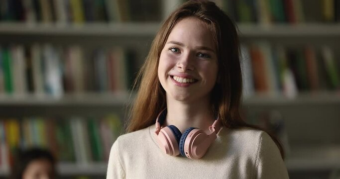 Close up portrait smiling pretty redhead student girl with wireless headphones looking at camera, posing in university or public library, head shot. Getting secondary or higher education, knowledge