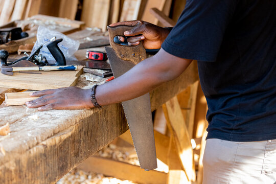 A young African carpenter saws wooden boards with a handsaw in a carpentry workshop.
