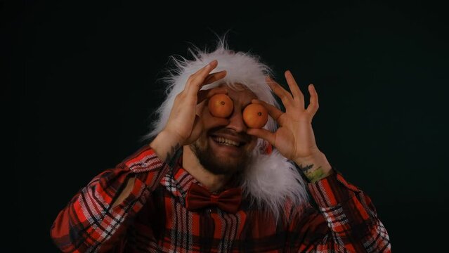 Caucasian young man with beard and tattoo wears red fluffy Santa hat and checkered shirt with bow tie. Man puts tangerines to eyes laughs against dark background. Happy New year and merry Christmas.