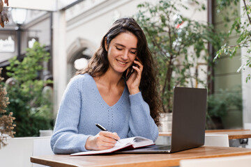 Happy girl is talking on the phone and chatting with colleagues remotely. Cheerful woman manager with laptop is making notes in notebook and talking with colleagues.