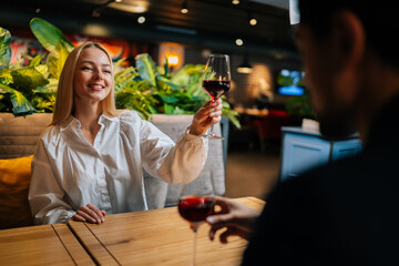 View from back of unrecognizable elegant man clinking glasses of red wine with cheerful pretty blonde woman sitting at table in fancy restaurant at evening. Happy couple enjoying nice romantic dinner.