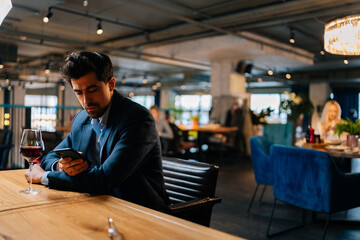 Pensive elegant young man in suit using smartphone chatting with friends and drinking red wine sitting at table in restaurant. Handsome bearded male waiting for girlfriend on date.