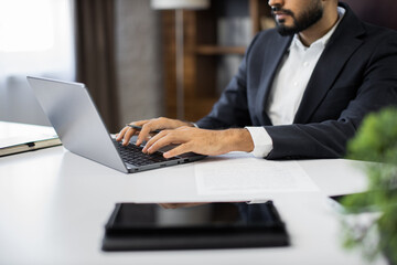 Fototapeta premium Cropped view of hands of young bearded businessman typing on laptop during work in office. Concentrated adult successful man wearing official suit sitting at wooden desk indoor.